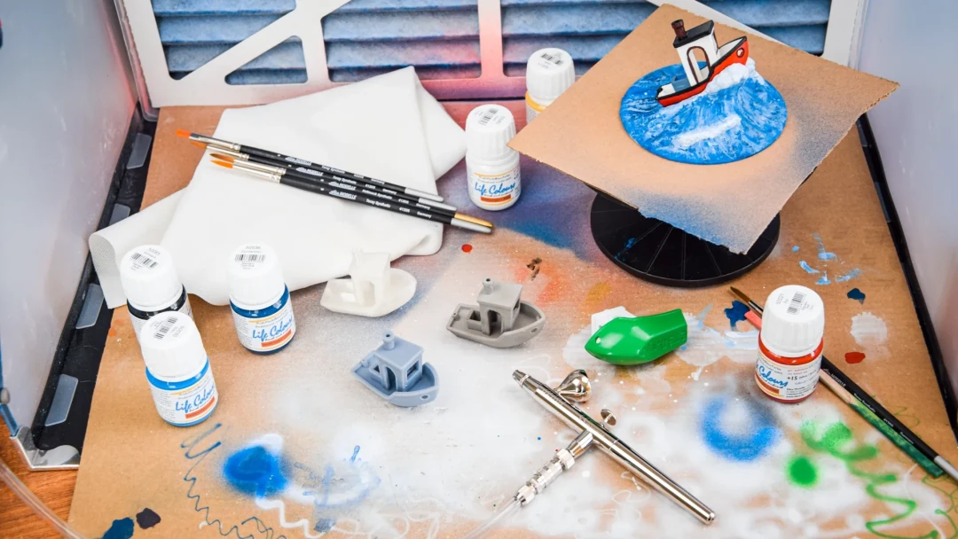 Ein Arbeitstisch in der Modell-Werkstatt, darauf verteilt kleine Schiffsmodelle, Pinsel, Airbrush und Modellbaufarben.